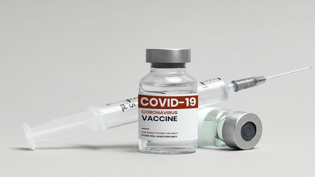 A COVID-19 vaccine vial and syringe on a clean white surface, with a blurred background showing medical equipment.