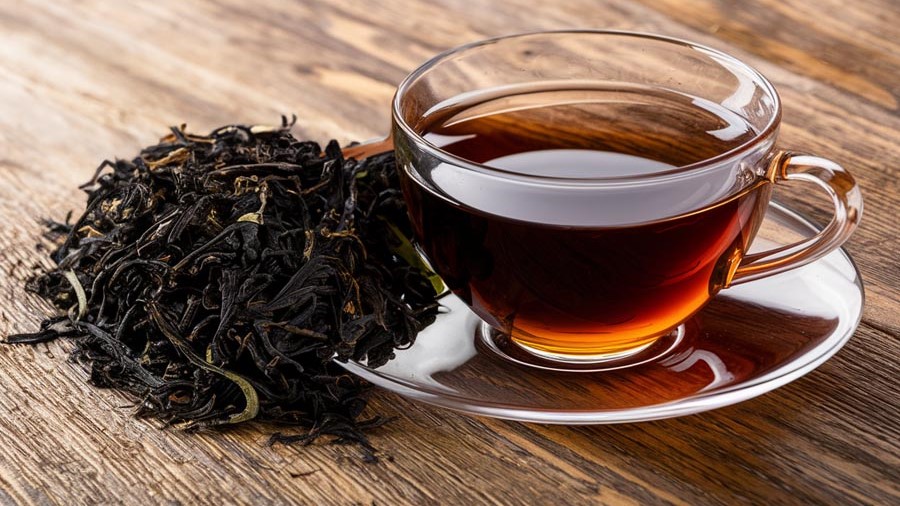 A steaming cup of black tea in a white ceramic mug, placed on a rustic wooden table with tea leaves and a slice of lemon beside it.