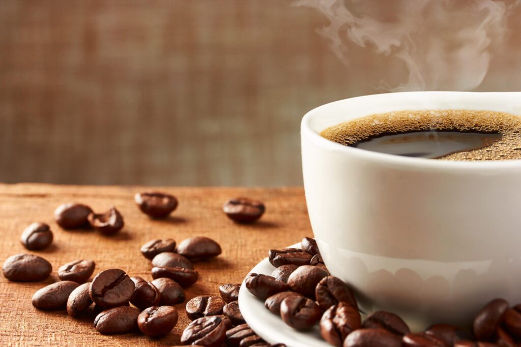 A cup of freshly brewed coffee on a wooden table, surrounded by roasted coffee beans, with steam rising from the surface of the drink.