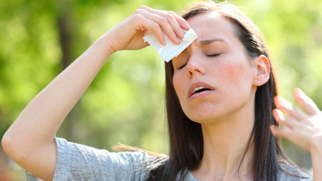 A person sitting outdoors under the sun, holding their forehead, with dry, cracked soil in the background symbolizing dehydration and extreme heat.