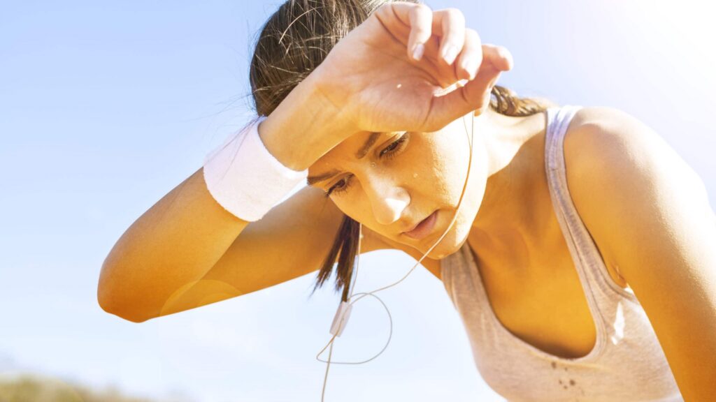 A person sitting outdoors under the sun, holding their forehead, with dry, cracked soil in the background symbolizing dehydration and extreme heat.