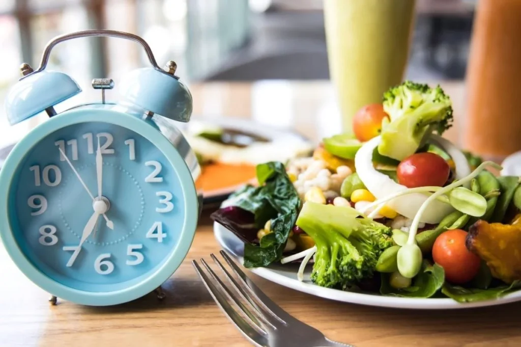 "Image of a clock surrounded by healthy foods such as fruits, vegetables, nuts, and a glass of water, symbolizing intermittent fasting and timed eating patterns."