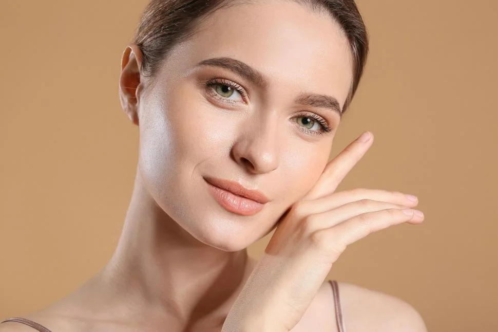 A young woman applying a moisturizing cream on her face, surrounded by various skincare products like serums, cleansers, and creams on a vanity table.